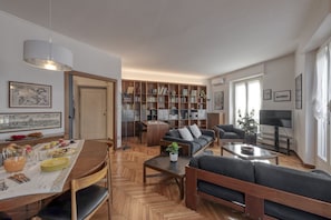 Ein modernes Wohnzimmer mit einer Couch, einem Couchtisch und einem Essbereich mit einem für das Frühstück gedecktem Tisch. Ein Bücherregal voller Bücher und ein Fernseher in der Ecke sind ebenfalls vorhanden.