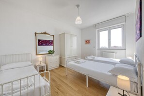 A hotel room with two beds, a white wardrobe, a small bedside table with a lamp, a window with a view, and a ceiling light.
