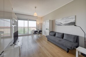 A modern living room with a grey sofa, a dining table with chairs, a TV, and a large window with a view.