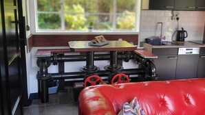 A modern kitchen with a red leather sofa, a window with a view of greenery, and a dining table with a kettle and a coffee maker.