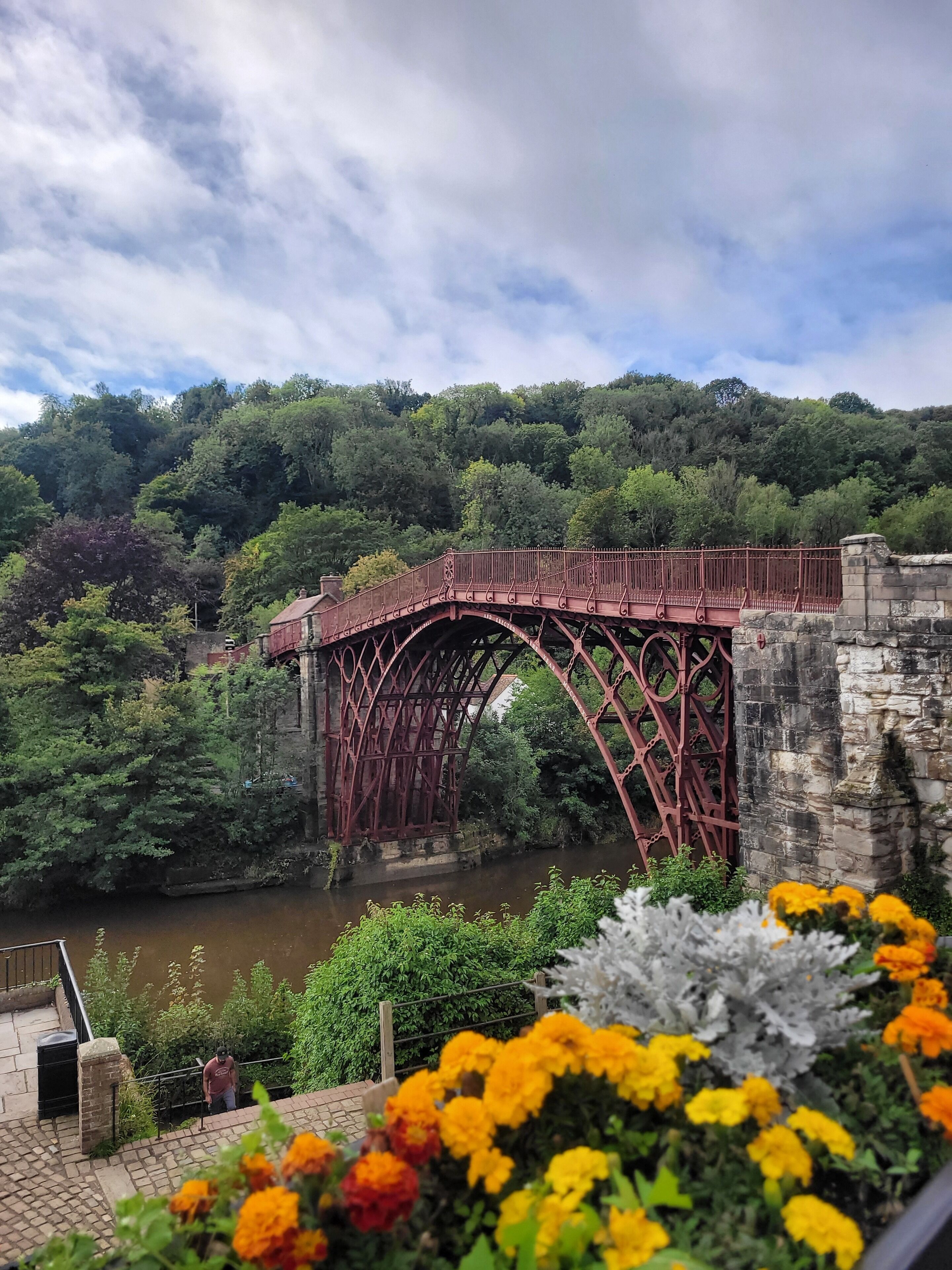 Un pont métallique rouge enjambant une rivière, entouré de verdure et bordé d’un mur de pierre.