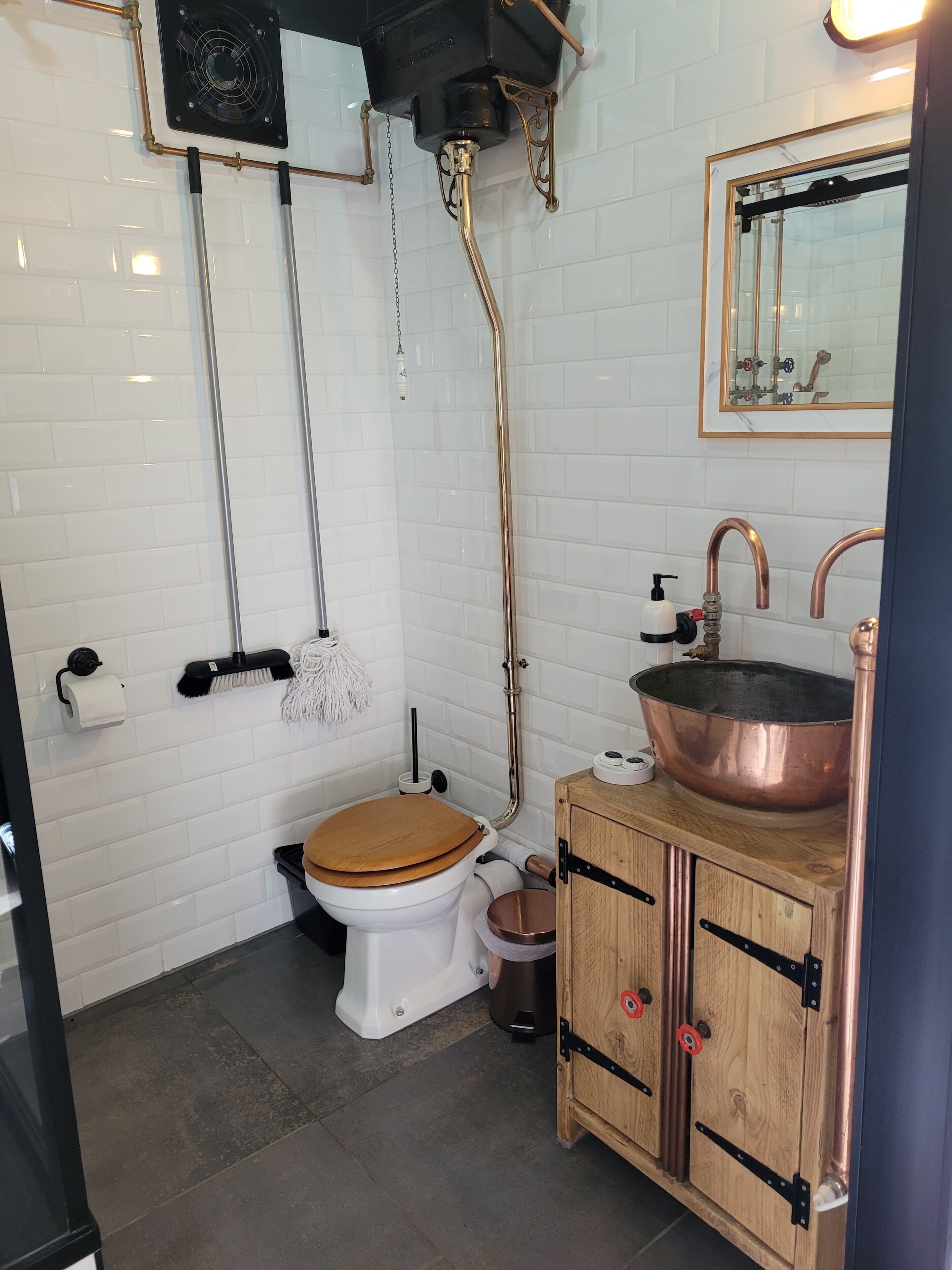 A modern bathroom with white tiled walls, a wooden toilet, a copper sink, and a wooden cabinet with metal handles.