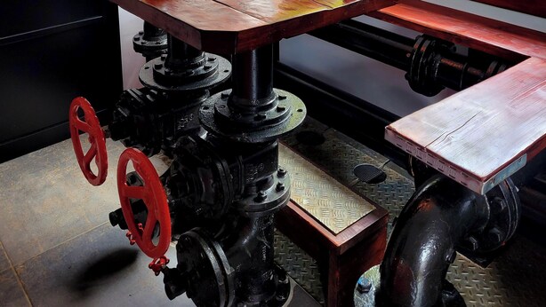 A dining area with a table set for two, a window with a view of greenery, and industrial-style pipes and wheels.