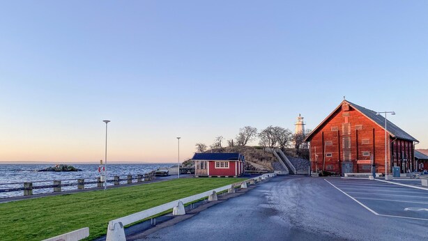 Una strada costiera con un fienile rosso, un piccolo edificio rosso e un faro sullo sfondo.