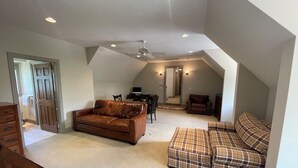 Une pièce mansardée spacieuse, avec un canapé en cuir marron, un divan à carreaux, une table à manger et des chaises, ainsi qu’un ventilateur de plafond.