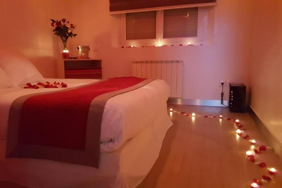 Quarto com cama, mesa de cabeceira com um vaso de flores e janela com vista para o exterior.