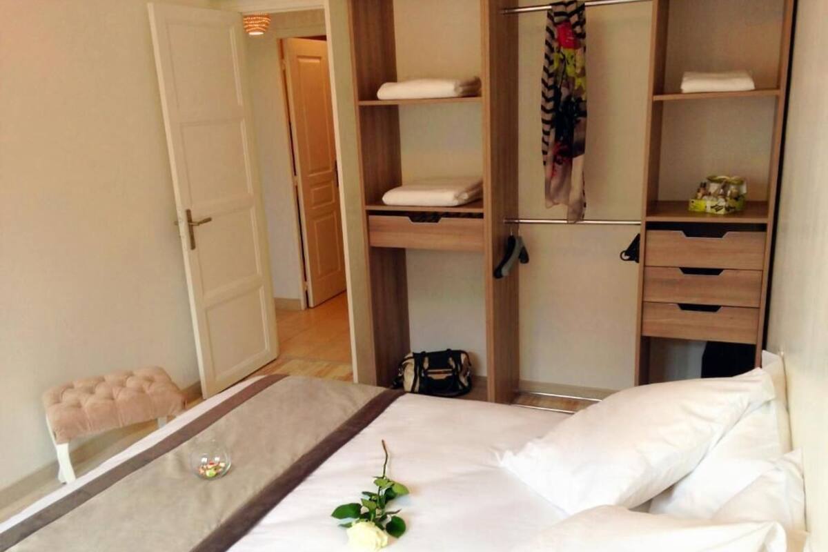 Cama bem arrumada com lençóis brancos, mesa de cabeceira com um copo, e um pequeno banco. Guarda-roupa de madeira com várias prateleiras e gavetas é visível.