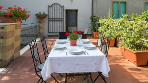 Une table dressée à l’extérieur, recouverte d’une nappe blanche et entourée de chaises, sur une terrasse carrelée agrémentée de plantes en pot, avec un bâtiment en arrière-plan.