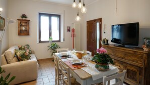 Un coin repas avec une table en bois dressée pour quatre personnes, entourée de chaises. Une télévision est posée sur un meuble en bois, à droite.