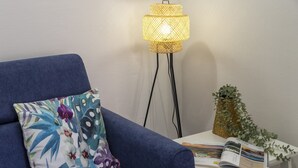 Un sofá azul con un cojín floral, una lámpara de pie, una mesita con un libro y una planta.