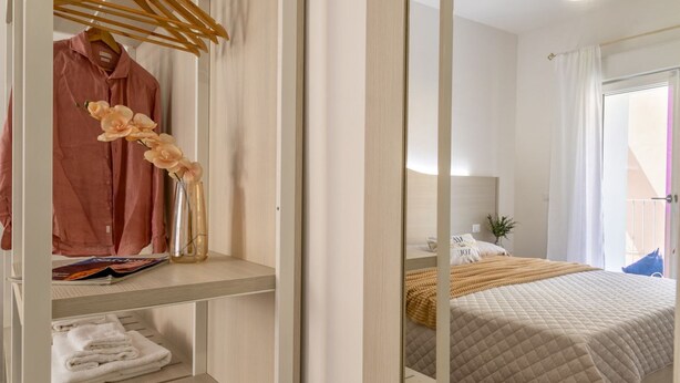 Un dormitorio con una cama, una mesita de noche, un espejo y un armario con ropa colgada y un jarrón de flores.