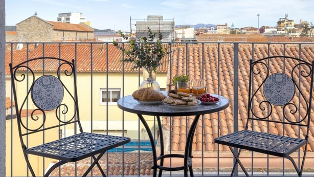 Un balcon avec deux chaises, une table garnie de nourriture et de boissons, et une vue sur des bâtiments en arrière-plan.