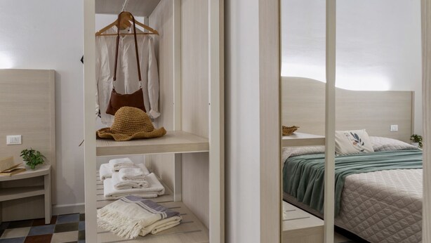 A modern bedroom with a bed, a shelf with folded towels, a hanging coat, and a small plant.