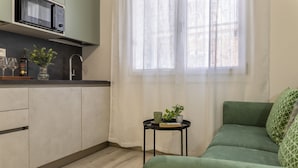 Una cocina moderna con microondas, copas de vino y un florero con flores. Hay un sofá verde y una mesita con una planta.