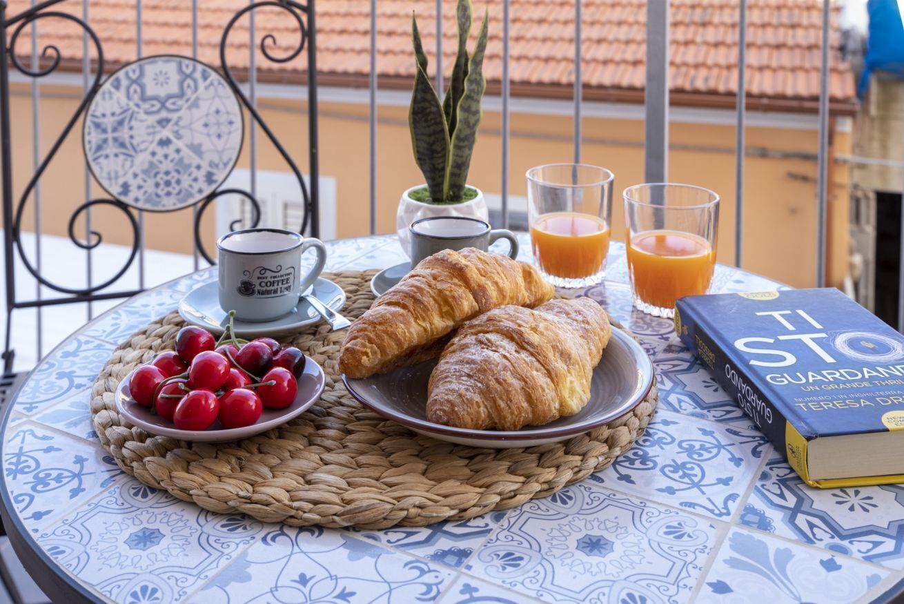 Ein Tisch mit Croissants, Kirschen, einem Buch, einer Kaffeetasse und Gläsern Orangensaft auf einer gemusterten Tischdecke.