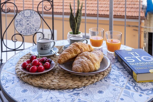 A table with croissants, cherries, a book, a coffee cup, and glasses of orange juice on a patterned tablecloth.