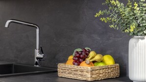 A kitchen counter with a woven fruit basket containing grapes, bananas, and apples, a modern faucet, and a white vase with green foliage. - Casa Erica. Apartment a few steps from the historic center. (Olbia)