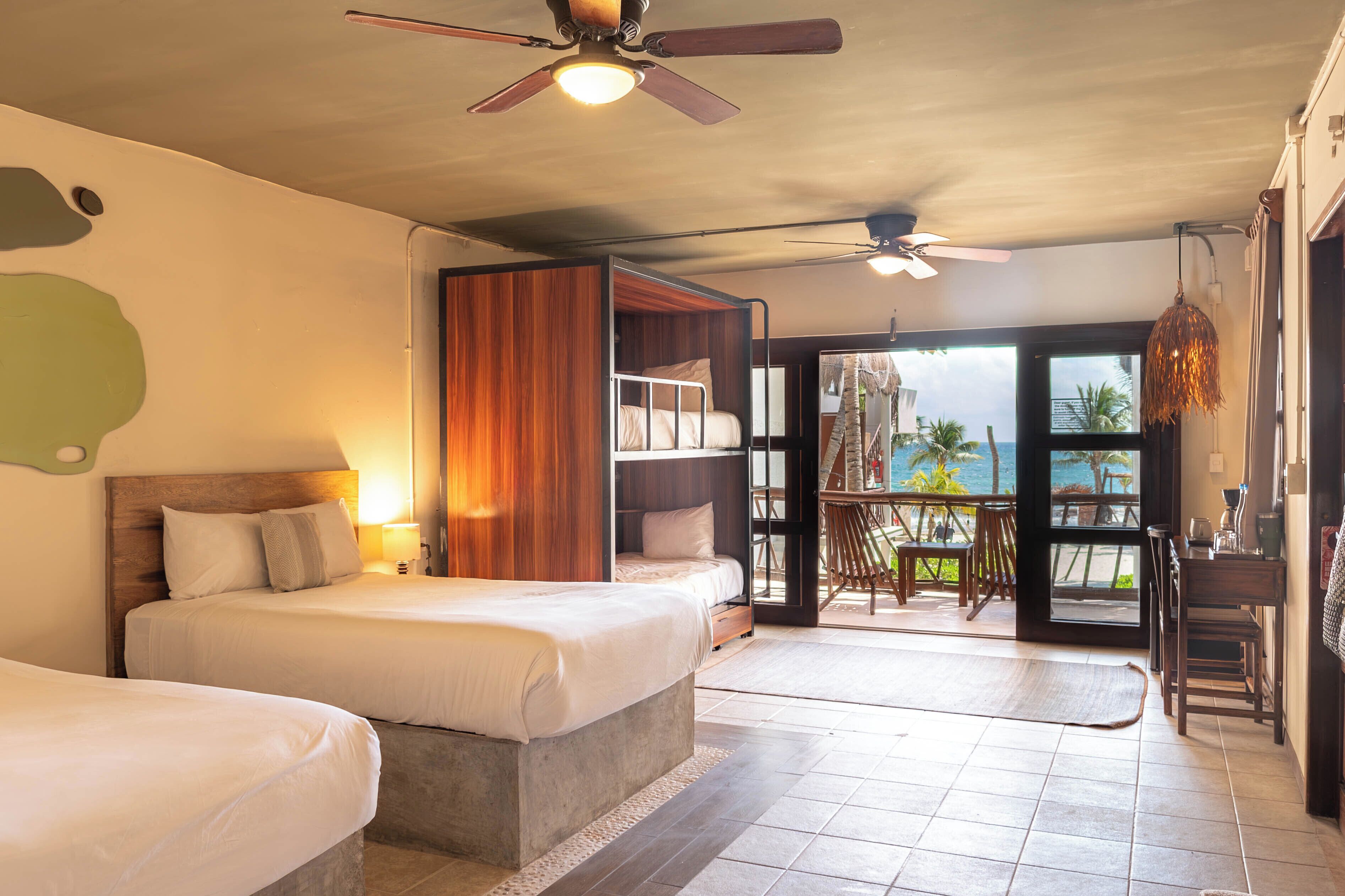 A hotel room with a bed, a ceiling fan, a balcony with a view of the ocean, and a dining area with chairs.