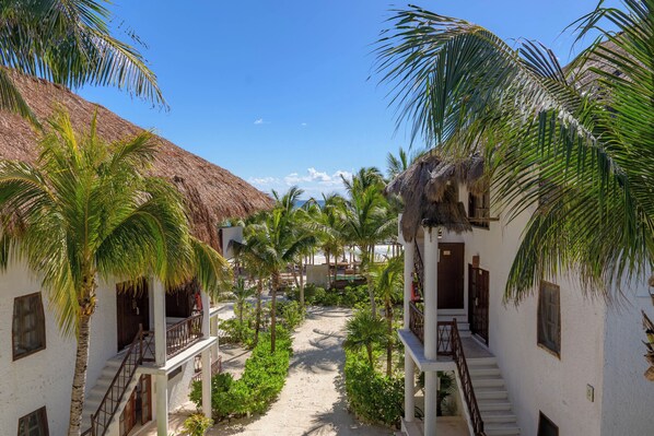 A pathway between buildings with palm trees, a beach in the background, and a clear sky. - Mía Tulum Beachfront Resort - Ocean View Suites and Beach Club (Tulum)