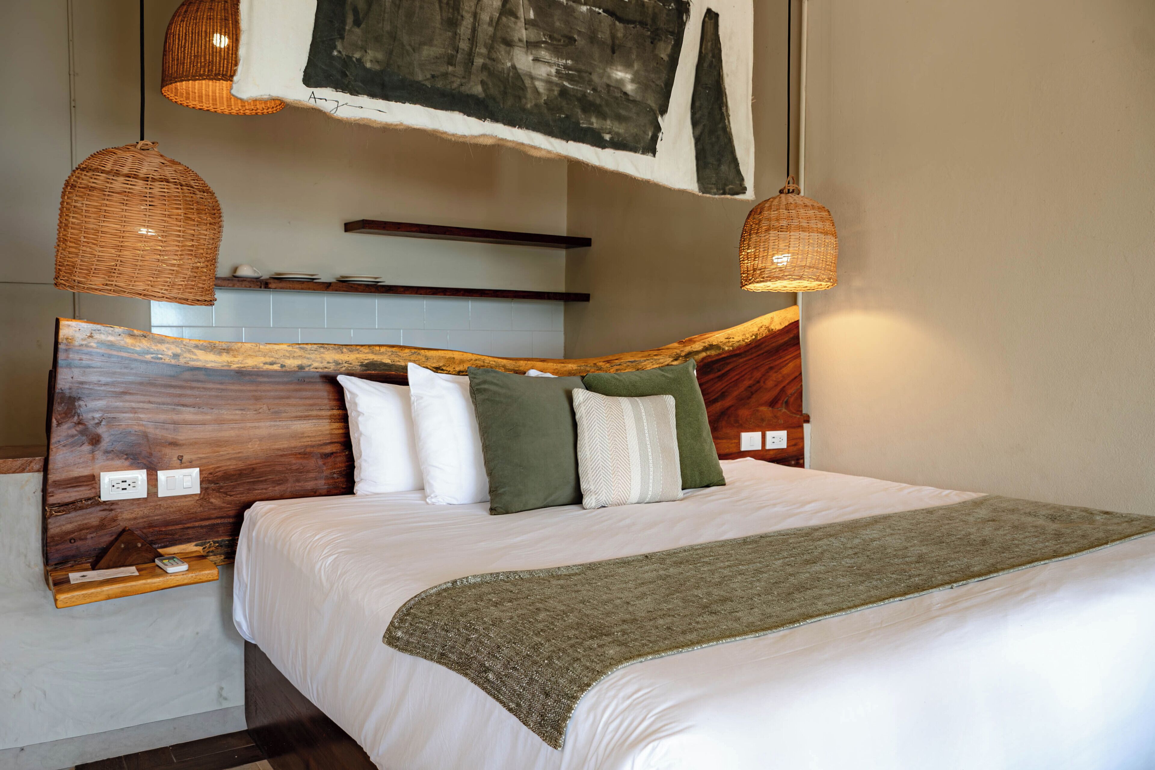 A bed with a wooden headboard, white bedding, and a green throw blanket. There are two wicker pendant lights above the bed.