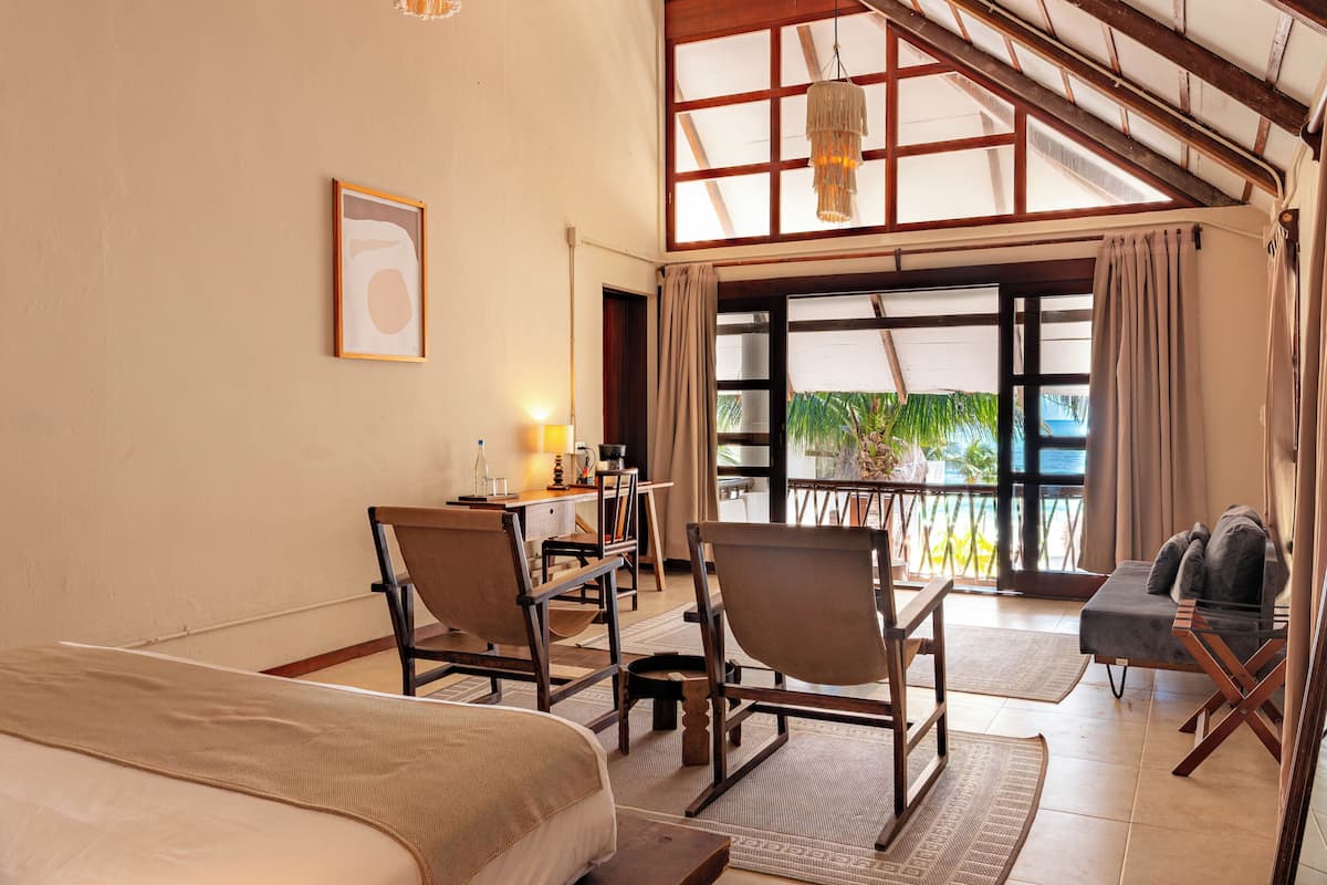 A room with a bed, a desk with chairs, a balcony with a view, and a hanging light fixture.