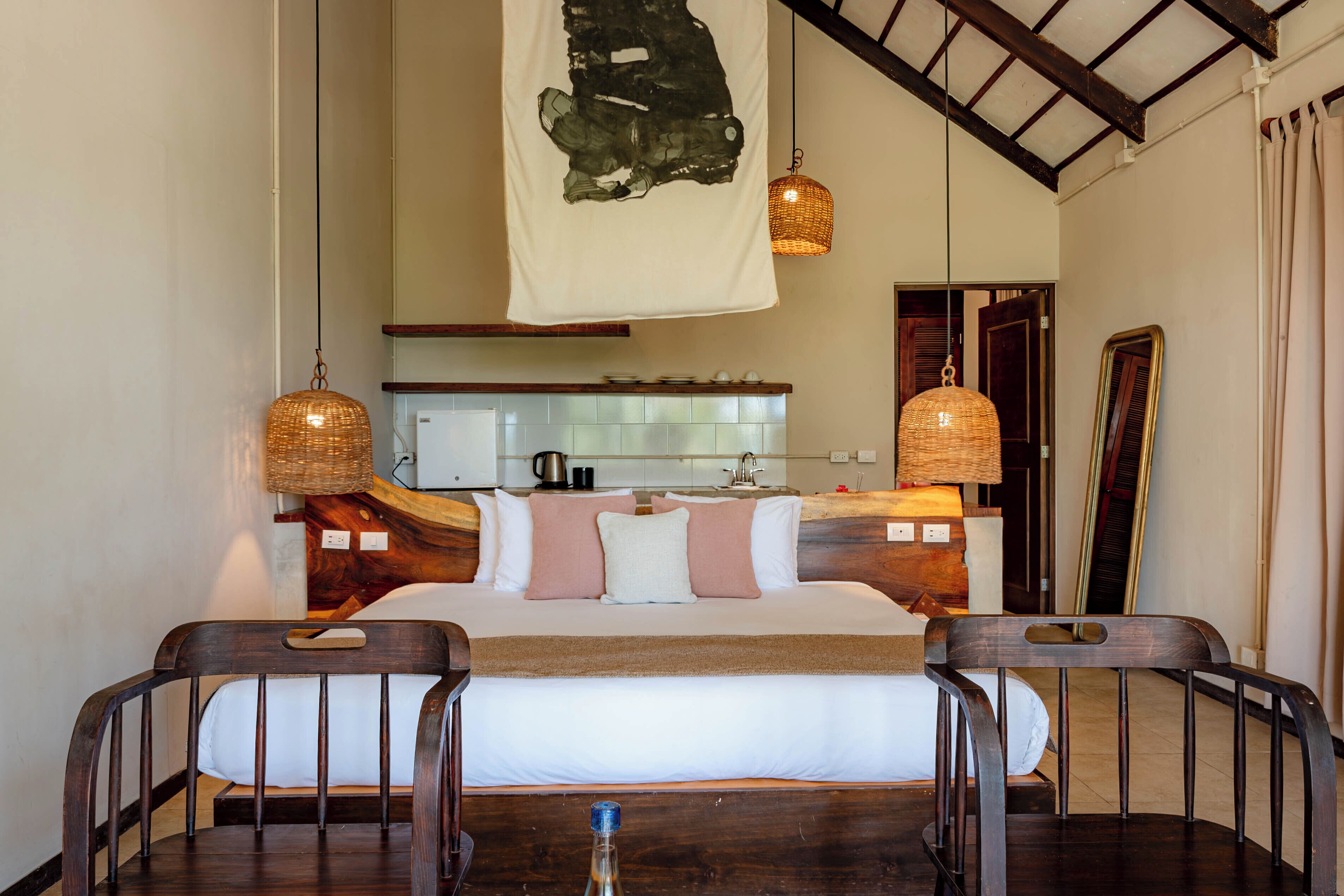 A hotel room with a bed, bedside tables, a hanging light fixture, a kitchen area, and a mirror.