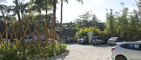 A sandy parking area with cars, palm trees, and a building with a thatched roof.