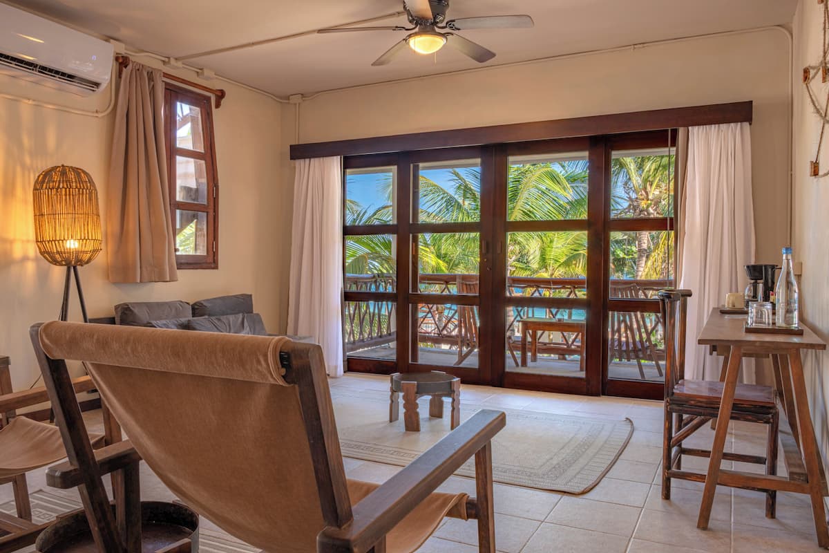 A room with a ceiling fan, a window with curtains, a wooden chair, a small table, and a view of palm trees outside.