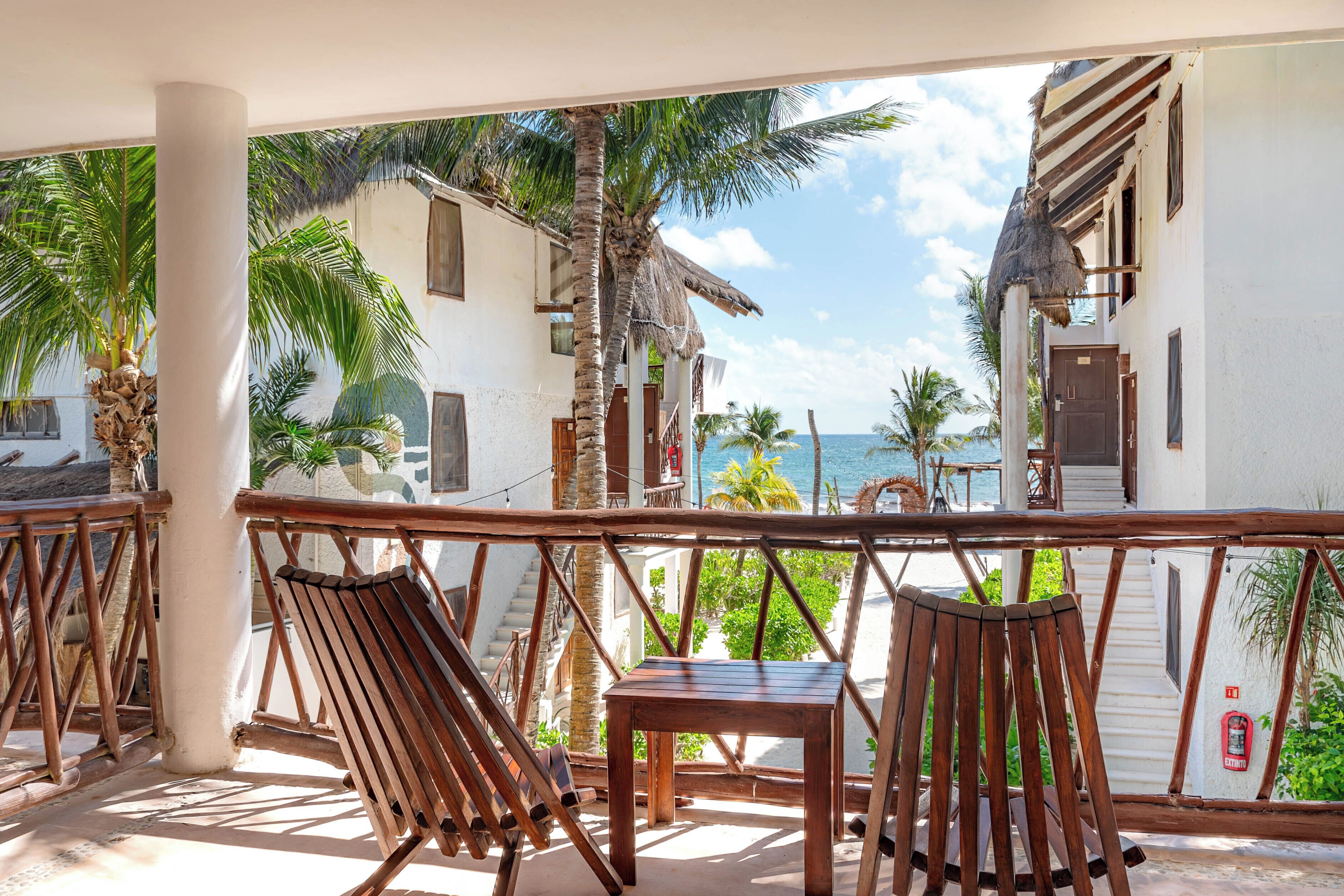 Ein Balkon mit Holzstühlen und einem Tisch, mit Blick auf einen Strand mit Palmen und ein Gebäude mit einem Reetdach.