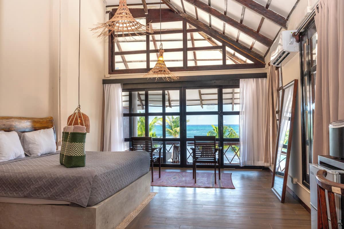 A bedroom with a large bed, a view of the ocean, and a balcony with wooden chairs.