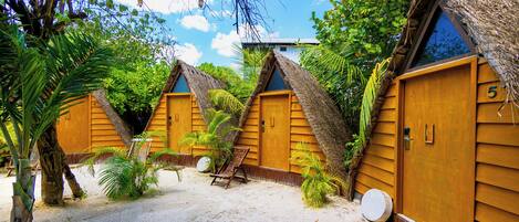 A row of wooden cabins with thatched roofs, numbered 5 and 6, surrounded by palm trees and sand.