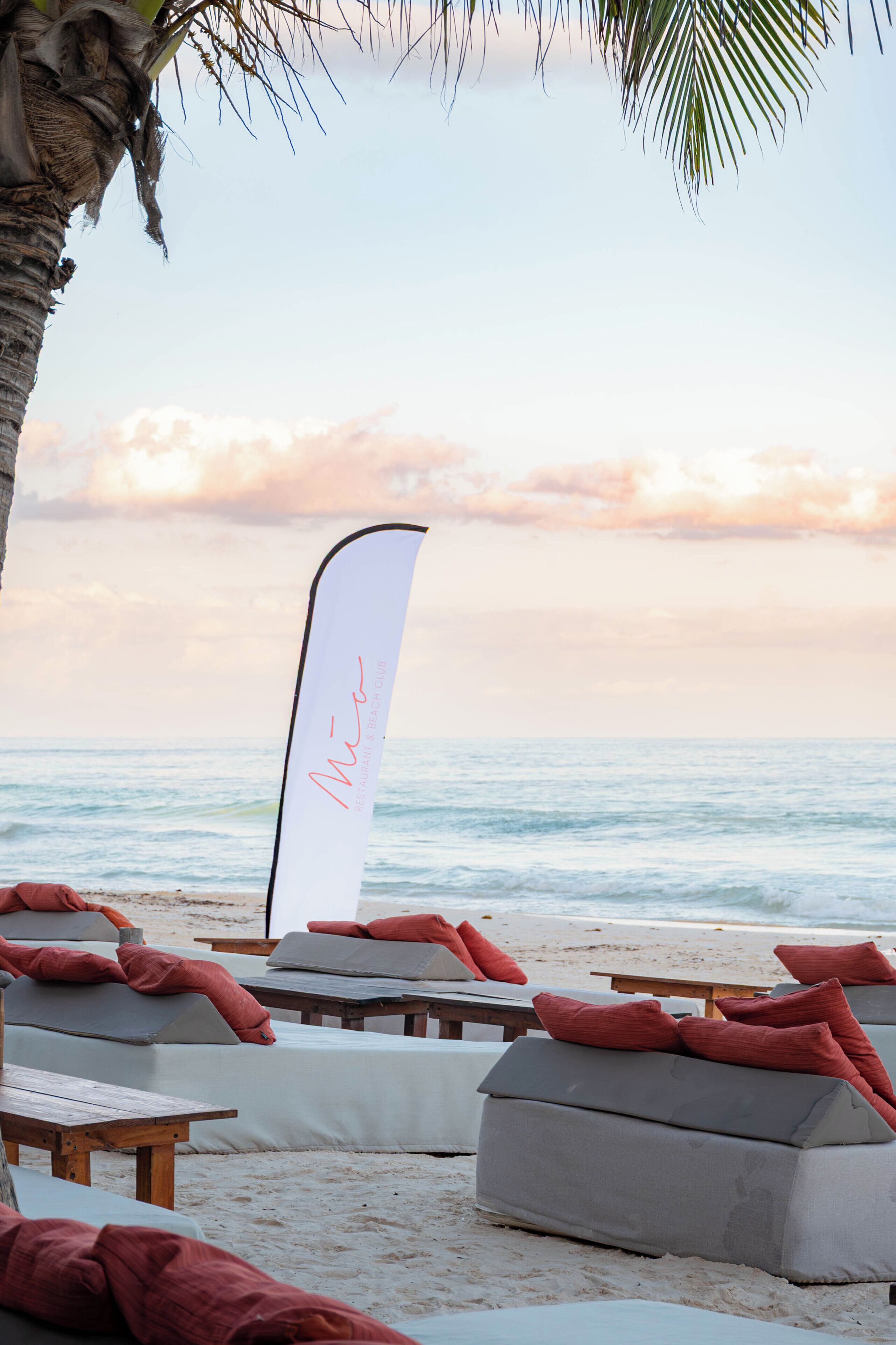 Un salón junto a la playa con asientos acolchados blancos y cojines rojos, y un banderín blanco con escritura cursiva.