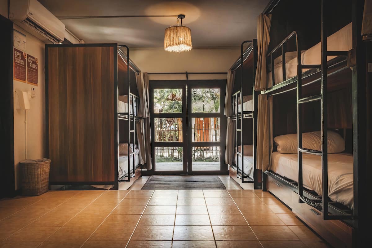 A hotel room with bunk beds, a wooden wardrobe, a woven light fixture, and a view through a glass door.