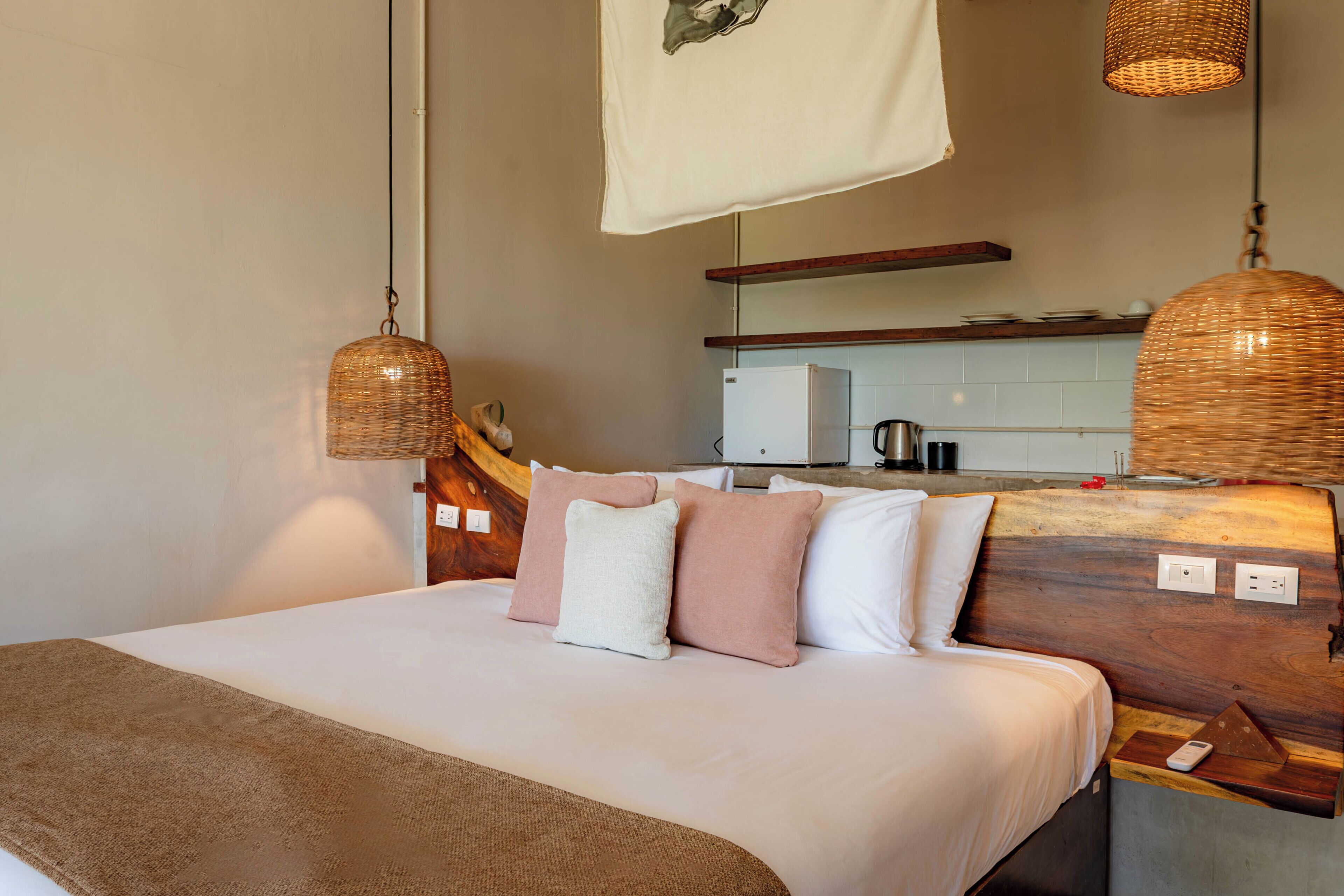 A bedroom with a bed, pillows, a bedside table, a lamp, and a kitchen in the background.