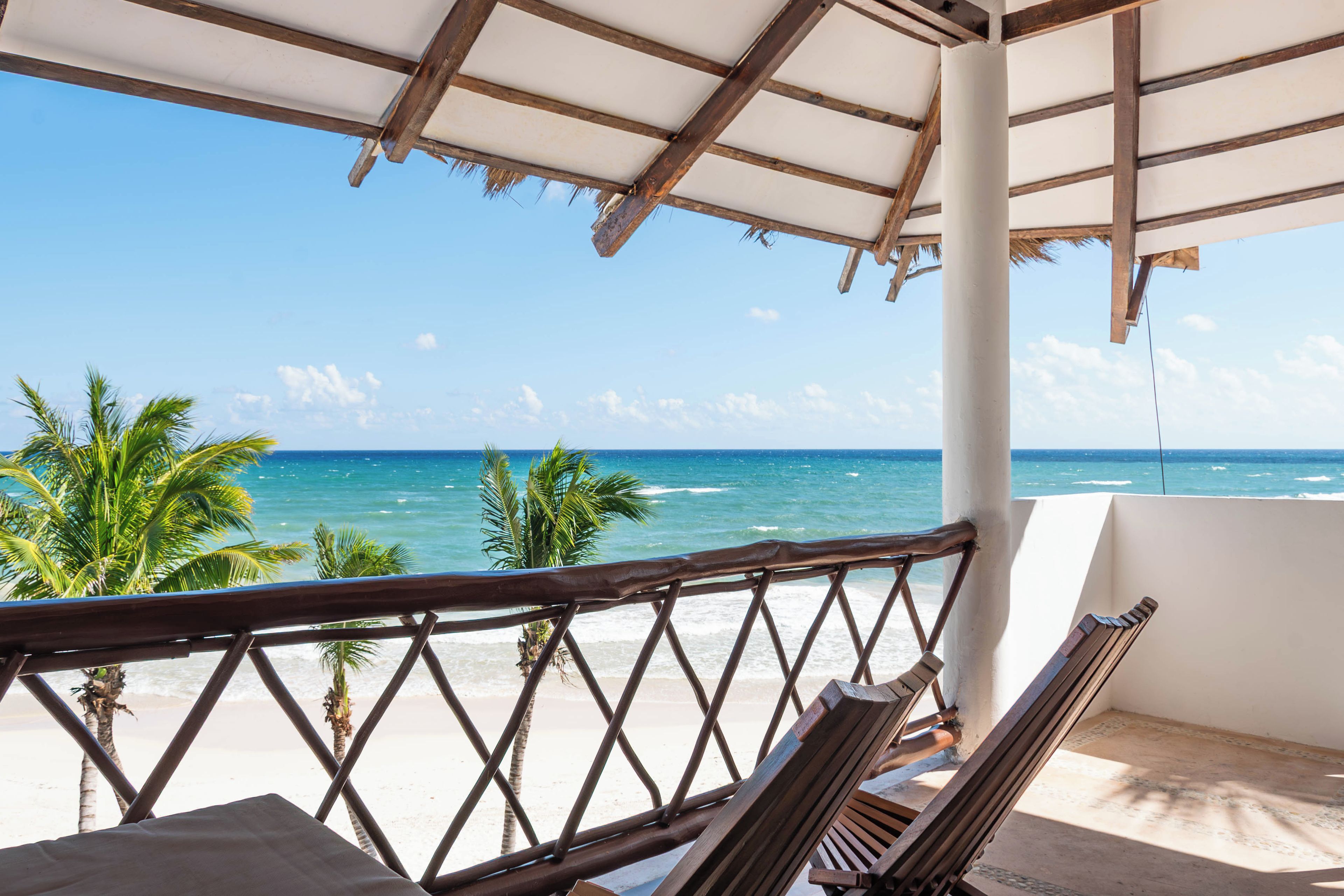 Vista a la playa desde un espacio cubierto con muebles de madera y palmeras.