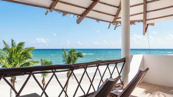 A beach view from a covered area with wooden furniture and palm trees.