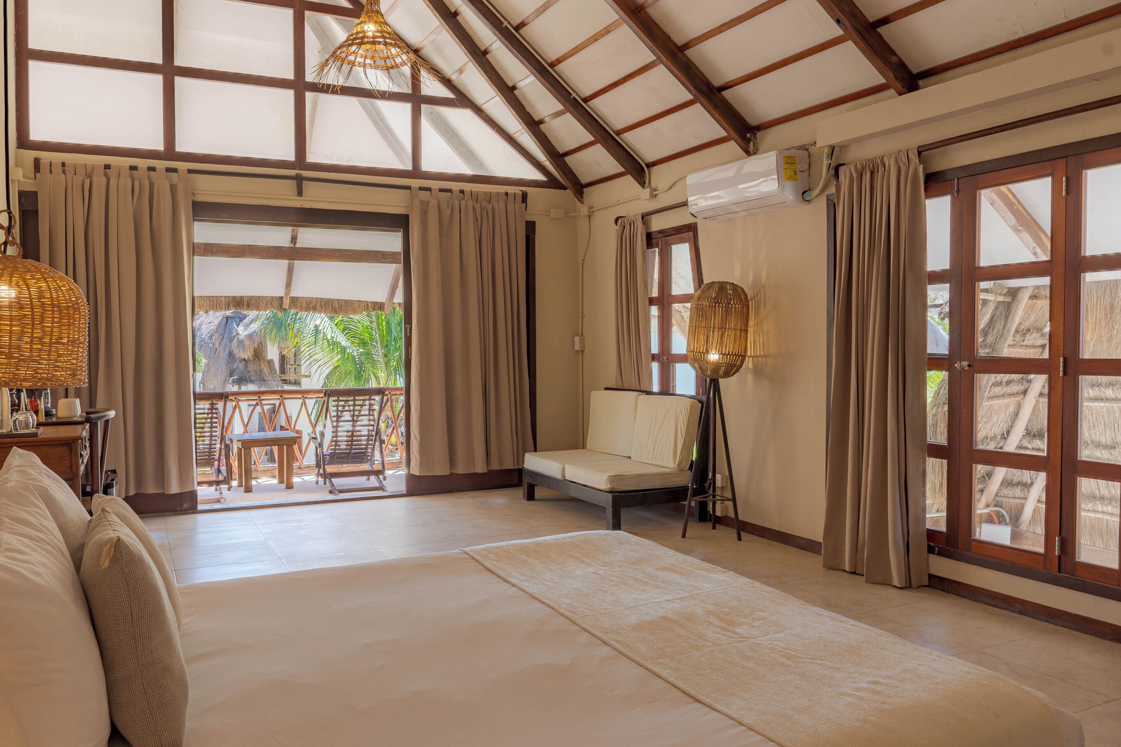 A spacious room with a large bed, a sofa, a small table, and a chair. There is a balcony with a view of palm trees.