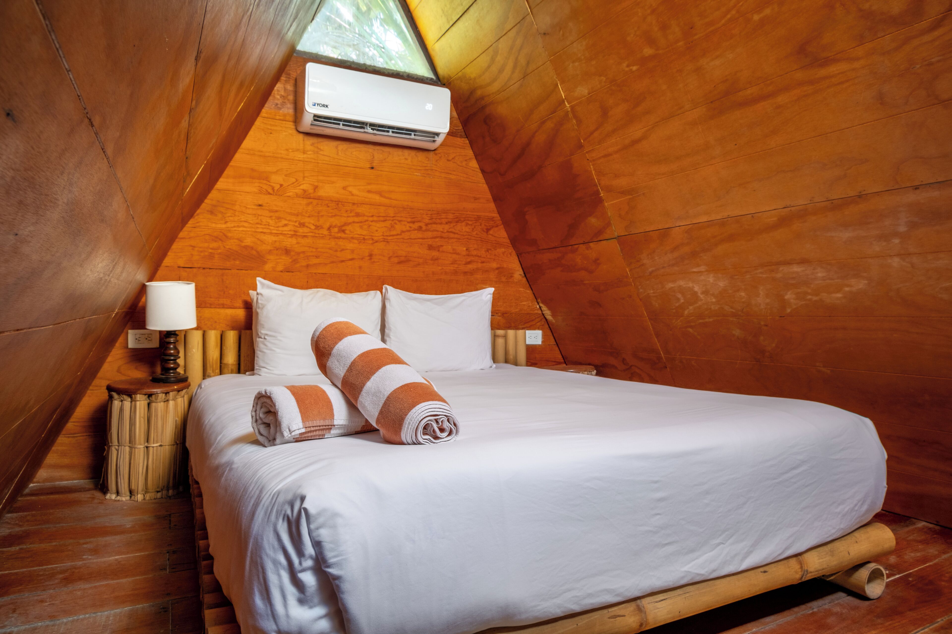 A bedroom with a bed, pillows, a rolled towel, a bedside lamp, and an air conditioning unit.
