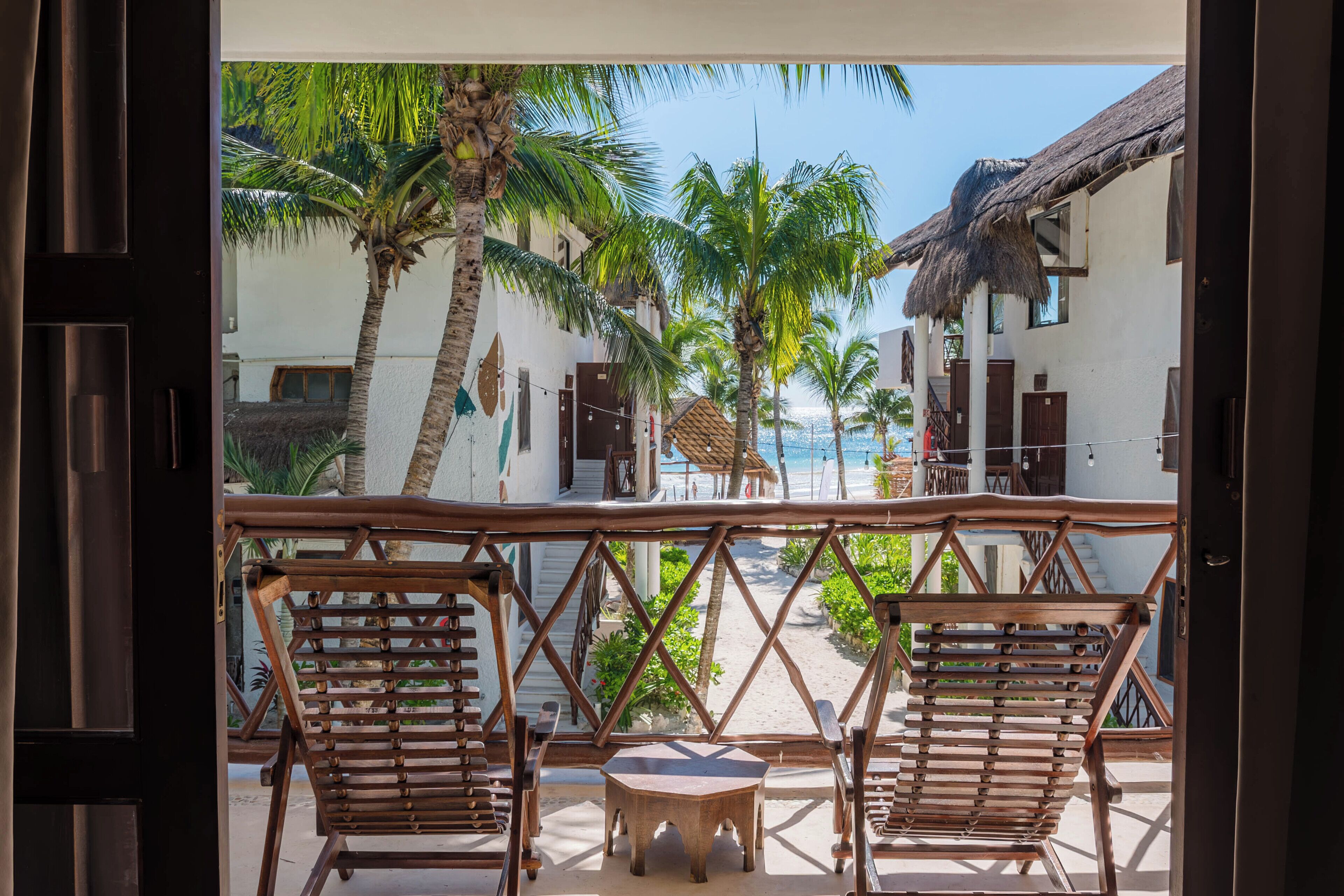 A balcony with two wooden chairs and a small table, overlooking a beach with palm trees and a building with a thatched roof.