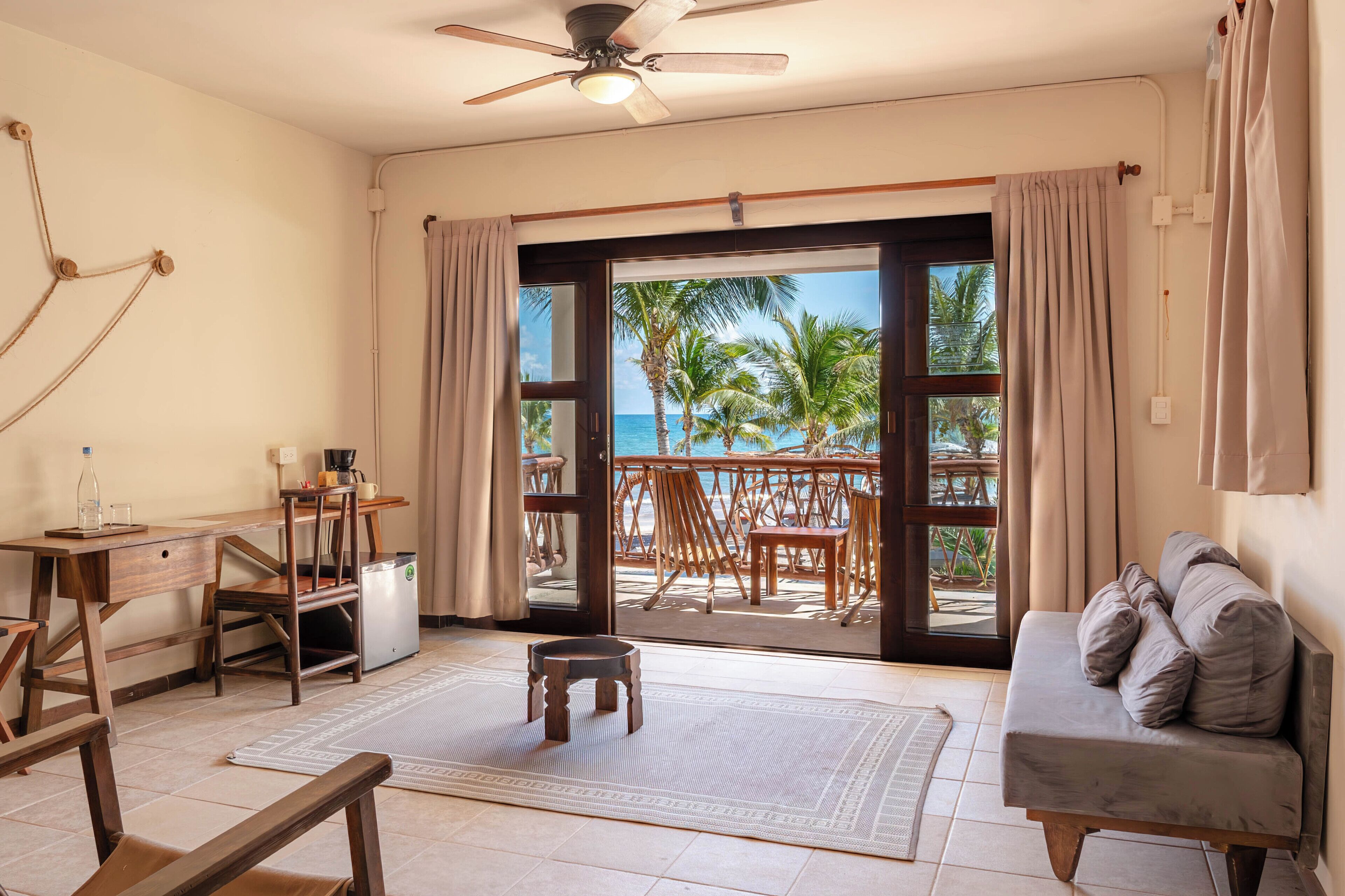 Een kamer met een bureau, stoel, bank en uitzicht op het strand door een grote schuifdeur.