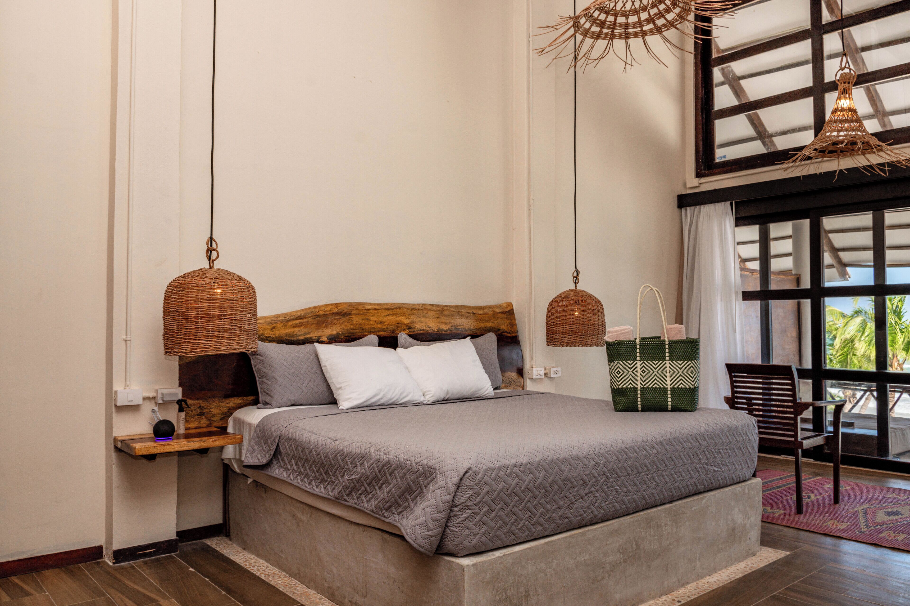 Un dormitorio con una cama grande, dos lámparas de mimbre colgantes, un cabecero de madera, una mesita de noche, un bolso tejido verde y vistas al exterior a través de un ventanal.
