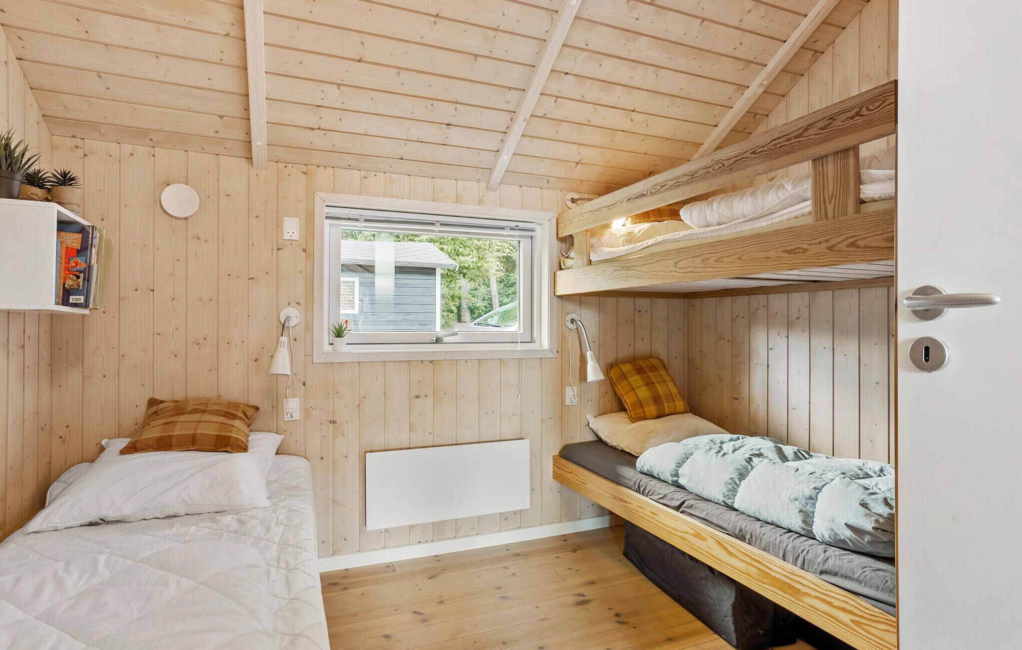 Ein gemütliches Blockhaus-Interieur mit Holzwänden und -böden, einem Etagenbett, einem Fenster mit Blick ins Grüne und einem Regal mit Büchern und Pflanzen.