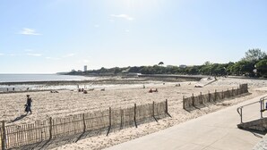 Una playa con una valla de madera, un camino de hormigón y una vista distante de edificios y árboles.