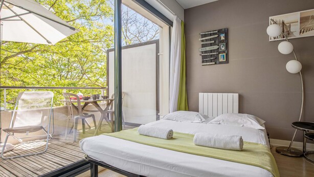 Un dormitorio moderno con una cama grande, un balcón con mesa y sillas, y vistas a árboles.