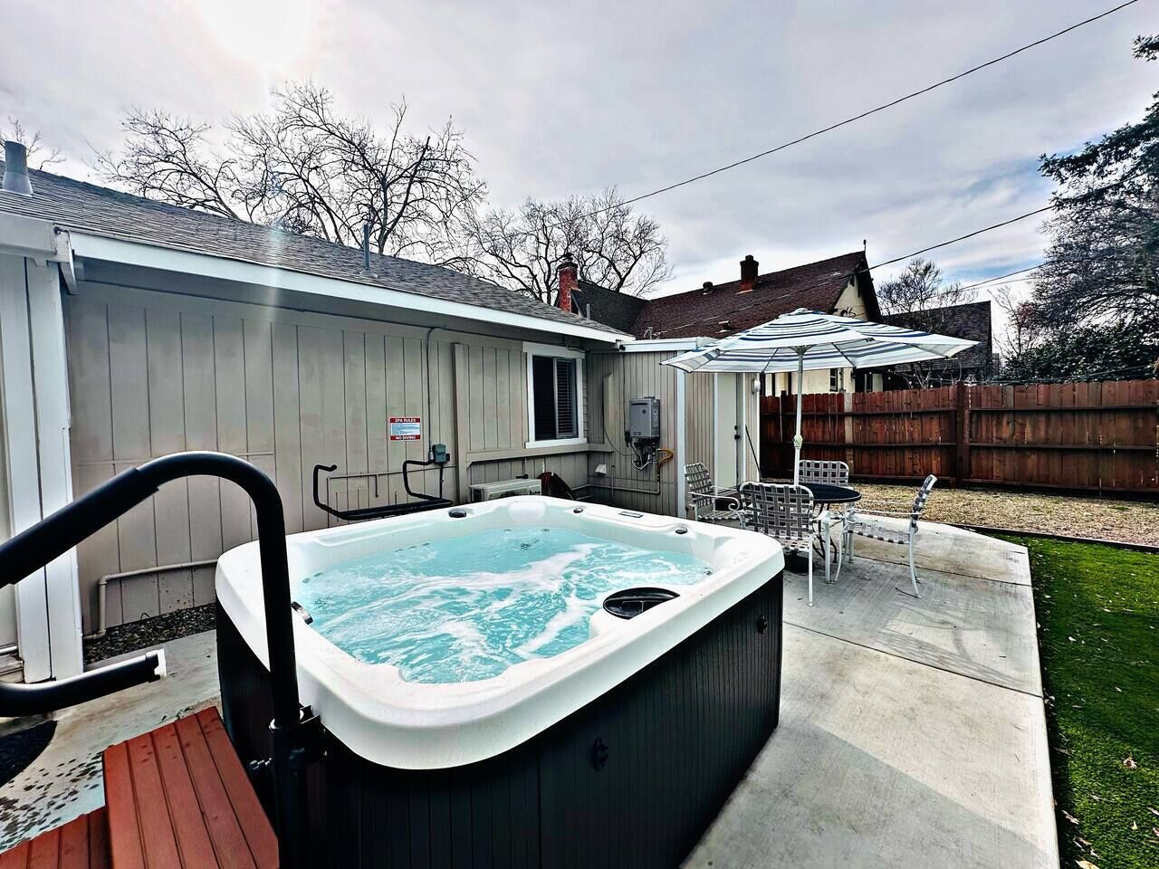 A hot tub in a backyard with a wooden deck, a patio table, and chairs.