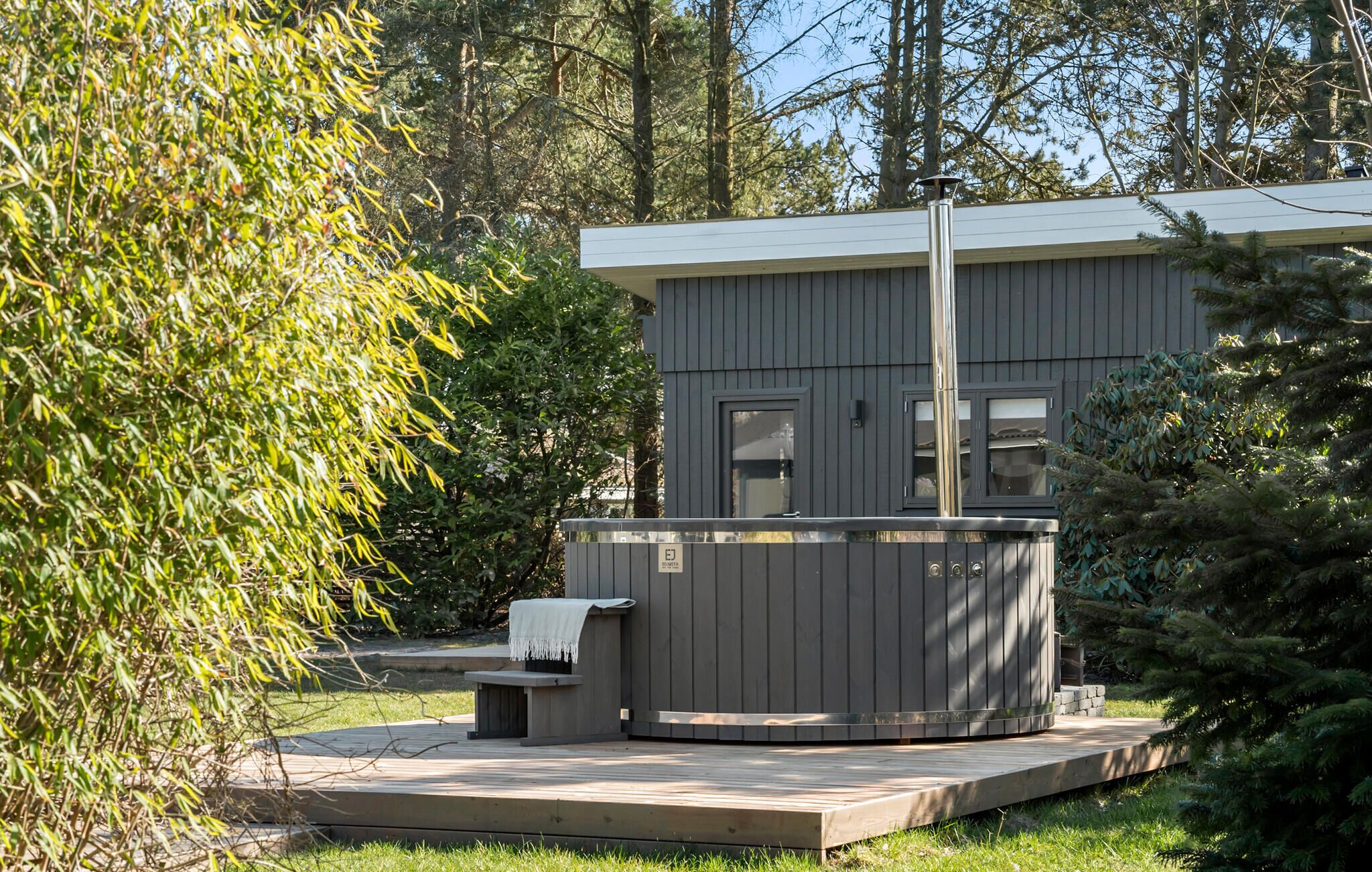 Un espace moderne aménagé à l’extérieur avec un bain à remous, doté d’une terrasse en bois, entouré d’une végétation luxuriante et avec un petit bâtiment en arrière-plan.