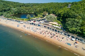 Une vue aérienne d’une plage bordée de rangées de parasols et de chaises longues, d’un parking et d’une route menant à un bâtiment dont le toit est recouvert de chaume.
