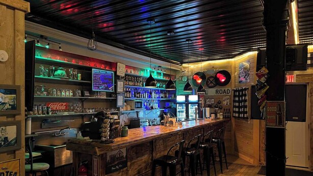 Un bar con scaffali in legno pieni di bottiglie, un neon e un bancone con sgabelli.