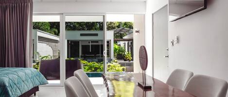 A modern hotel room with a bed, a dining table, chairs, a ceiling fan, and a view of a swimming pool and greenery outside.