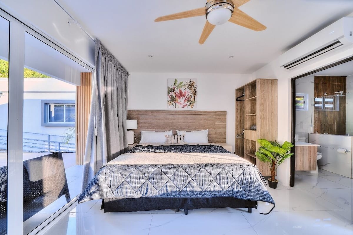 Quarto moderno com cama grande, ventilador de teto, planta em vaso e vista para o exterior.